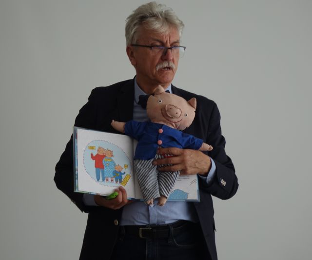 pisarz pokazuje książkę i maskotkę prosiaczka w niebieskim pulowerku