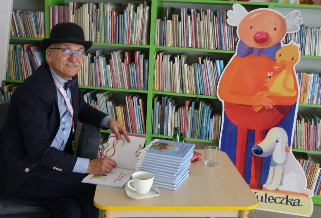 pisarz siedzi przy stoliku i podpisuje swoje książki, w tle regały z książkami