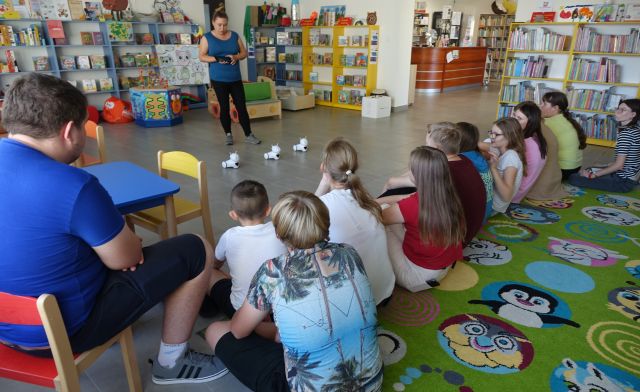 młodzież siedzi na kolorowym dywanie, przed nimi stoi bibliotekarka i omawia jak sterować robotami Photon