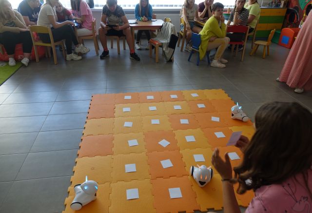 młodzież siedzi przy stolikach, dziewczyna kuca przed pomarańczową matą i steruje robotami Photon