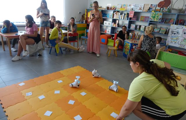 dziewczyna patrzy na roboty na pomarańczowej macie, przed nią przy stolikach siedzi młodzież