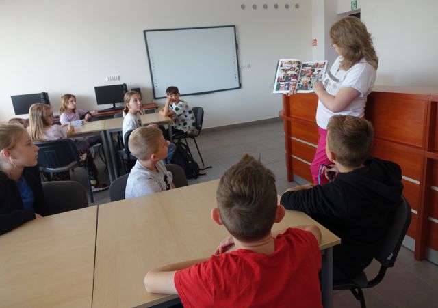 czwartoklasiści w sali komputerowej siedzą przy stołach, przed nimi stoi bibliotekarka i pokazuje komiks
