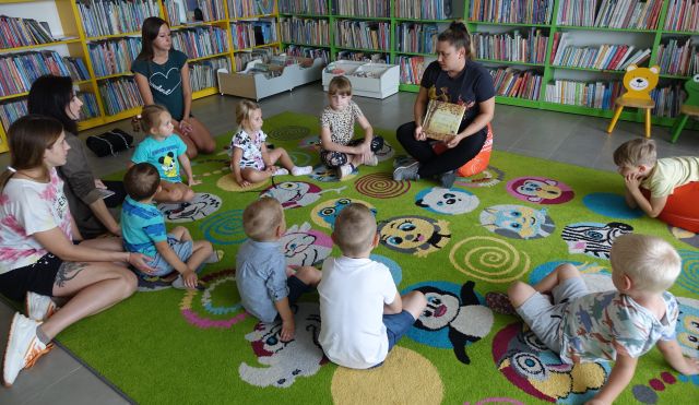 dzieci i rodzice siedzą na kolorowym dywanie, przed nimi siedzi bibliotekarka i pokazuje im książkę