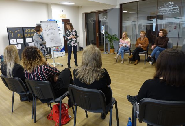 13 bibliotekarek siedzi w okręgu na krzesłach, przed nimi przy flipcharcie stoi kobieta