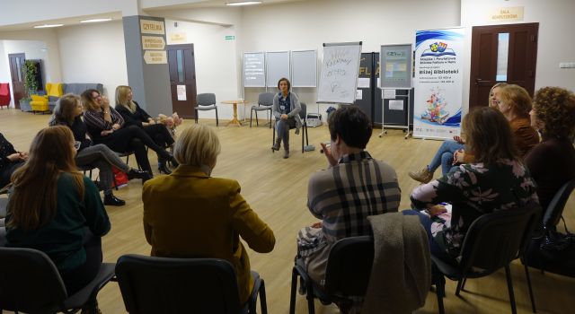 14 kobiet siedzi na krzesłach w kole, przed nimi siedzi kobieta