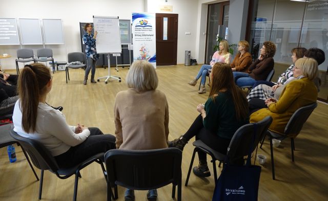kobieta pokazuje na flipchart, 15 kobiet siedzi na krzesłąch w kole