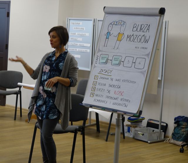kobieta stoi obok zapisanego flipcharta i mówi do uczestników szkolenia