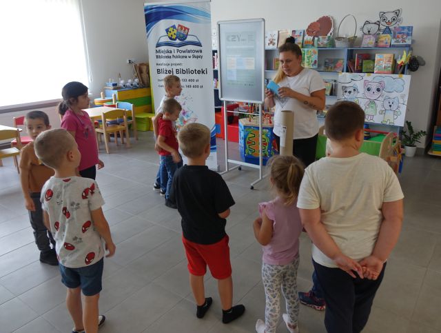 dzieci i bibliotekarka stoją, ze nimi banery z nazwą biblioteki i projektu oraz regały