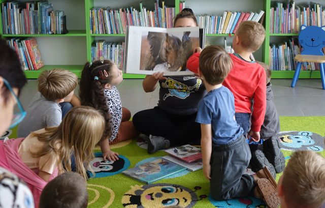 bibliotekarka pokazuje przedszkolakom książkę o zwierzętach, w tle regały z książkami