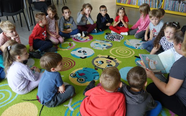 przedszkolaki siedzące na kolorowym dywanie słuchają jak bibliotekarka czyta im książkę