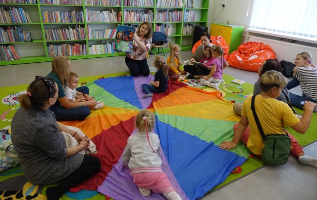 mali i duzi uczestnicy klubu malucha siedzą na kolorowym dywanie, bibliotekarka pokazuje książkę