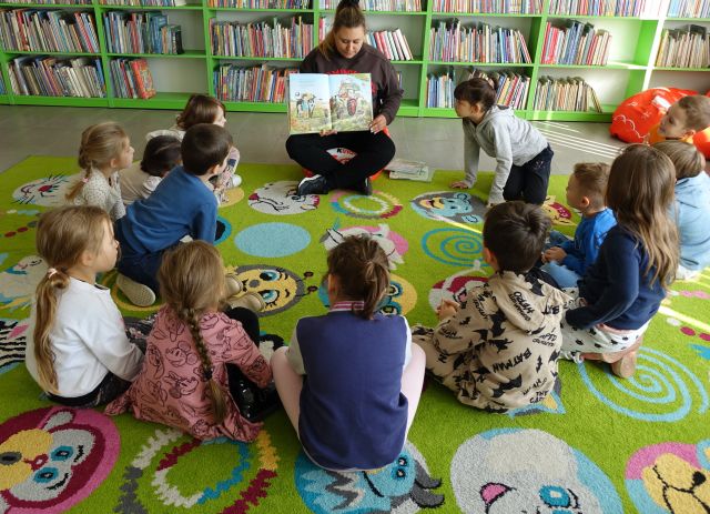 przedszkolaki siedzą na kolorowym dywanie, przed nimi siedzi kobieta z książką, w tle regały z książkami