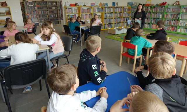 dzieci siedzą przy stolikach, przed nimi stoi bibliotekarka, w tle regały z książkami