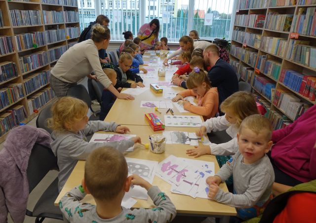 dzieci i opiekunowie wykonują prace plastyczne siedząc przy stołach