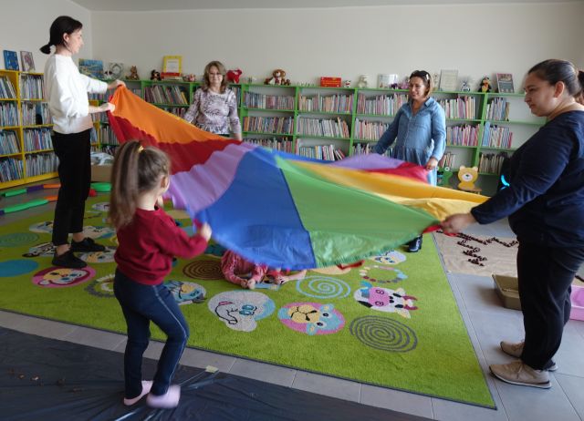 4 dorosłe kobiety i chłopiec trzymają kolorową chustę animacyjną, pod chustą na kolorowym dywanie leżą dzieci