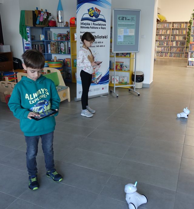 chłopiec i dziewczynka sterują robotami Photon przy użyciu tabletów, w tle regały i banery z nazwą projektu i biblioteki