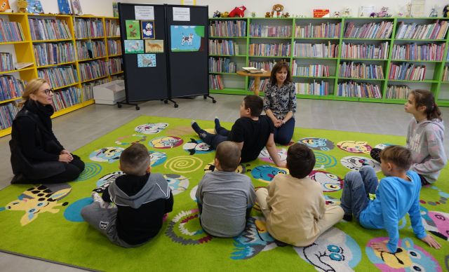 dzieci i dwie osoby dorosłe aiwdzą na kolorowym dywanie, w tle regały z książkami