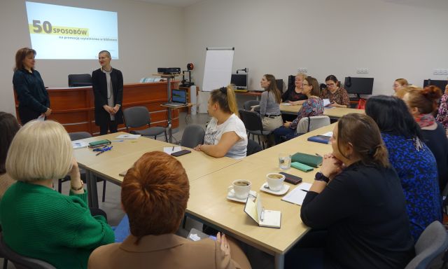 uczestnicy szkolenia siedzą przy stołach ustawionych w podkowę, w tle biurka z komputerami