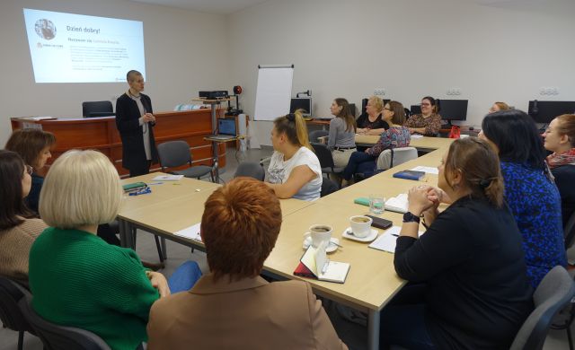 uczestnicy szkolenia siedzą przy stołach ustawionych w podkowę, w tle biurka z komputerami