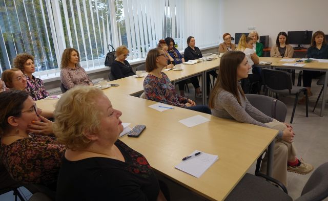 uczestnicy szkolenia siedzą przy stołach ustawionych w podkowę, w tle biurka z komputerami