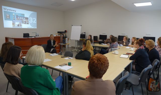 uczestnicy szkolenia siedzą przy stołach ustawionych w podkowę, w tle biurka z komputerami