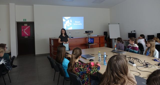 młodzież siedząca przy stołąch słucha bibliotekarki, w tle sali komouterowej prezentacja wyświetlona na ścianie