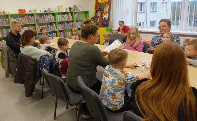 dzieci i rodzice siedzący przy stole słuchają czytającej bibliotekarki