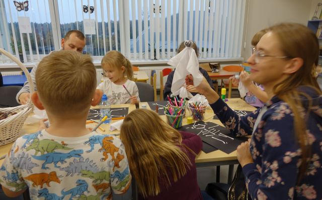 dzieci siedzą przy stołąch i wykonują ludziki z patyczków oraz lalki z białęgo materiału