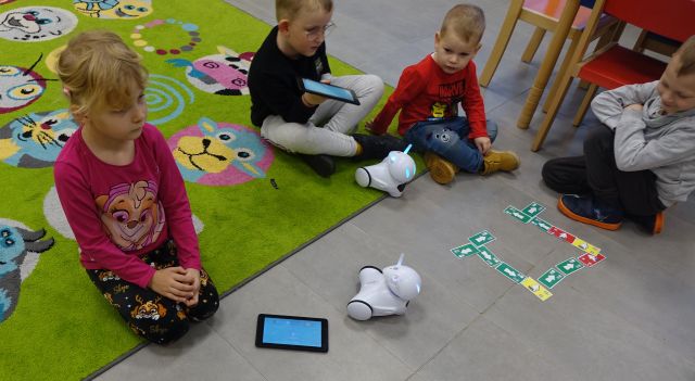 dzieci siedzą na kolorowym dywanie i sterują przy pomocy tabletów robotami Photon jeżdżącymi po karteczkach