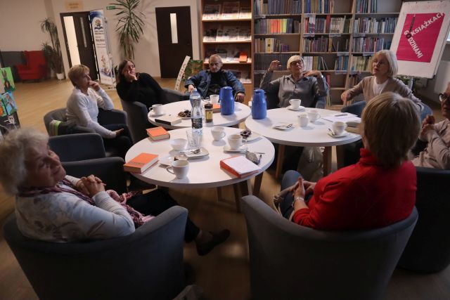 uczestnicy Dyskusyjnego Klubu Książki siedzą przy okrągłych stołach, na których mają książki, filiżanki i termosy