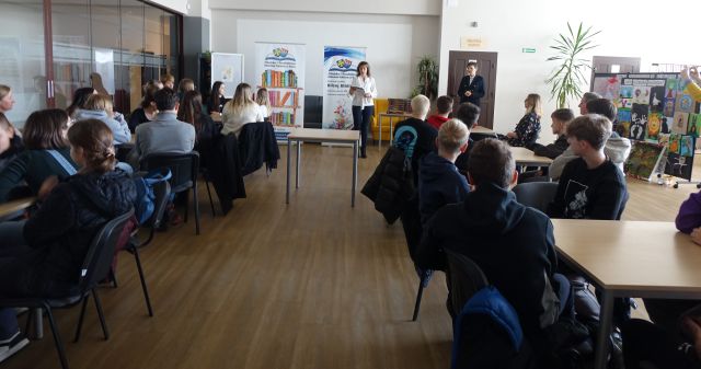 młodzież siedząca przy stołach, przed nimi w tle dwie kobiety oraz baner biblioteki i z nazwą projektu