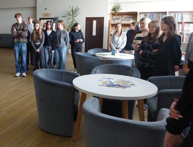 młodzież w czytelni biblioteki, stoi pomiędzy siwymi fotelami i białymi ogrągłymi stołami