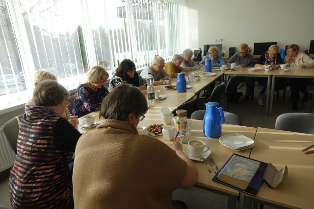 seniorki siedzą przy stołach z kawą i ciastkami i piszą na kartkach