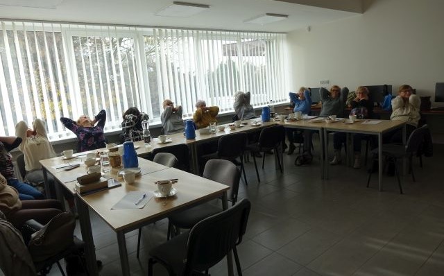 Seniorki siedząc na krzesłach przy stolikach wykonują ćwiczenia