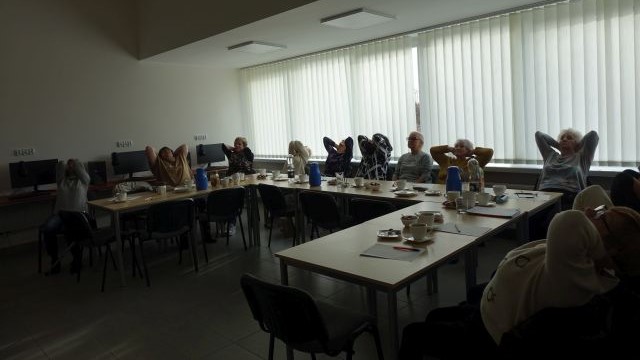 Seniorki siedząc na krzesłach przy stolikach wykonują ćwiczenia