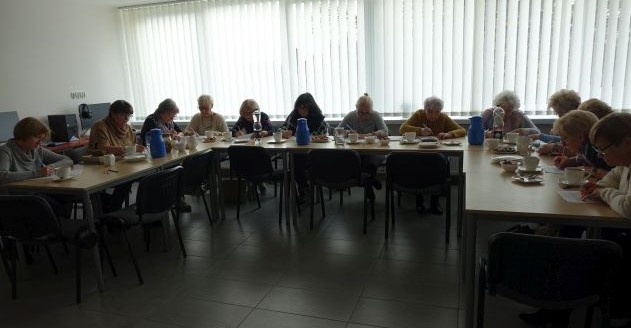 Seniorki siedząc na krzesłach przy stolikach wykonują ćwiczenia