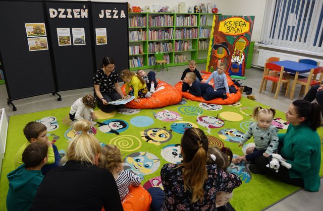 dzieci i opiekunowie siedzą na kolorowym dywanie i słuchają czytanej przez bibliotekarkę książki, w tle regały z książkami i czarna tablica z napisem dzień jeża i ilustracjami jeży