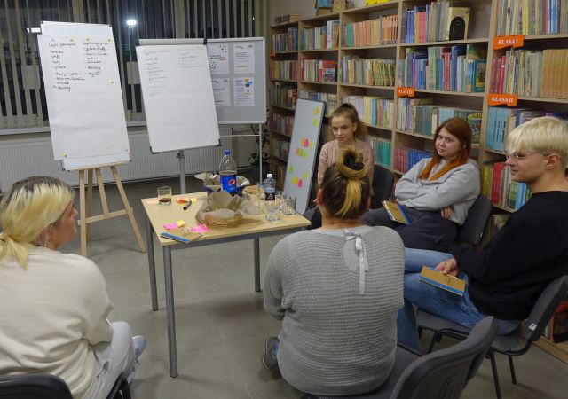 młodzież siedzi na krzesłach, przed nimi stól z chipsami i napojami oraz flipchart, w tle regały z książkami