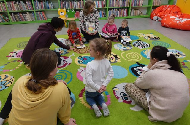 dzieci i rodzice siedzą na kolorowym dywanie i przeglądają książki
