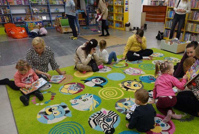 dzieci i rodzice siedzą na kolorowym dywanie i przeglądają książki
