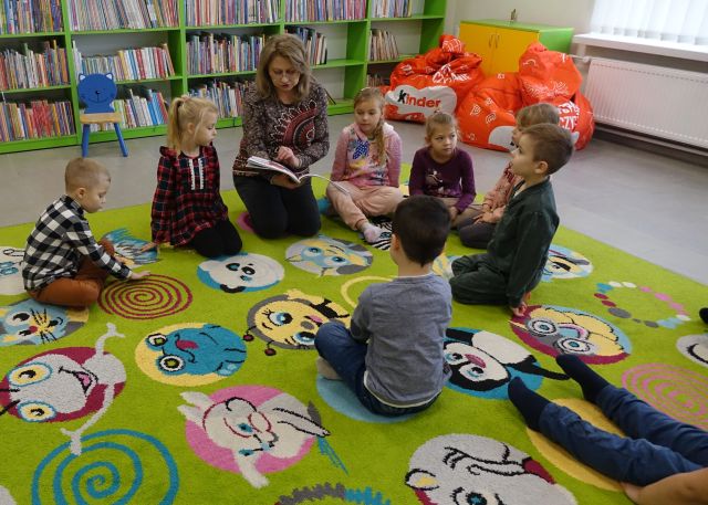 przedszkolaki siedzą na kolorowym dywanie i słuchają książkę którą czyta im bibliotekarka