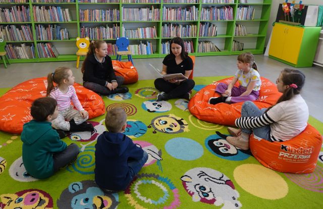 dzieci i bibliotekartka z książką siedzą na kolorowym dywanie, w tle regały z książkami