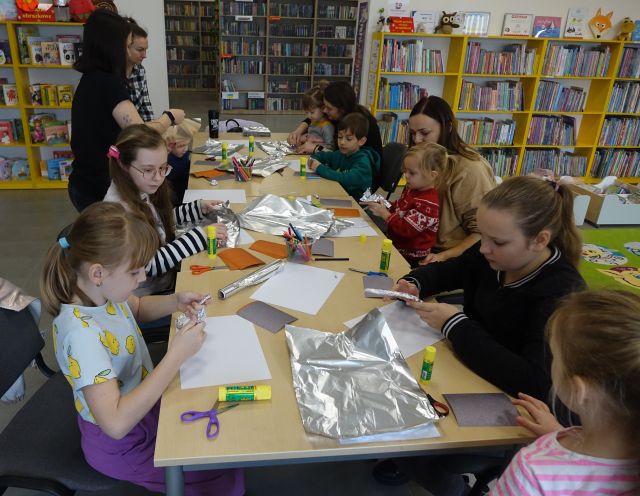 dzieci i rodzice siedzą przy stołach i wykonują prace z folii aluminiowej