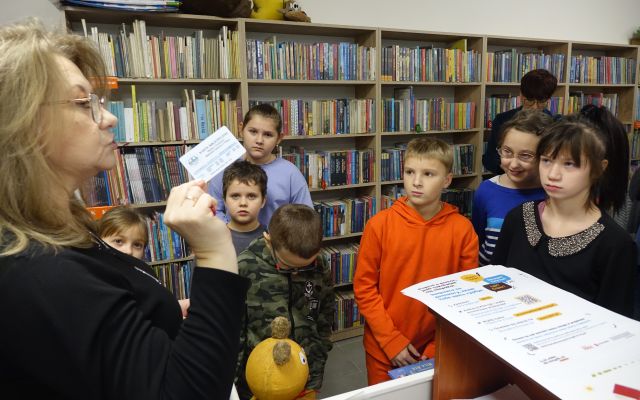 dzieci przed bibliotekarką z kartą biblioteczną w dłoni, w tle regały z książkami
