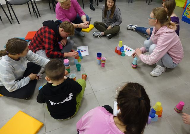 dzieci i młodzież siedzą na podłodze i ukłądają kolorowe kubeczki