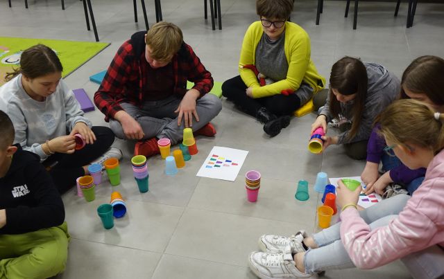 dzieci i młodzież siedzą na podłodze i ukłądają kolorowe kubeczki