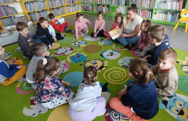 przedszkolaki siedzą na kolorowym dywanie, siedząca między nimi bibliotekarka pokazuje im książkę