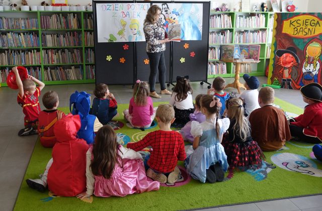 dzieci w przebraniach za postacie z bajek siedzą na kolorowym dywanie, przed nimi stoi kobieta z książką