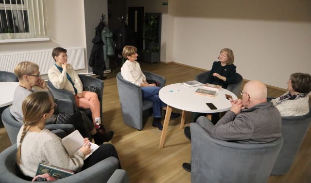 6 kobiet i 1 mężczyzna siedzą na siwych fotelach przy białych okrągłych stołach