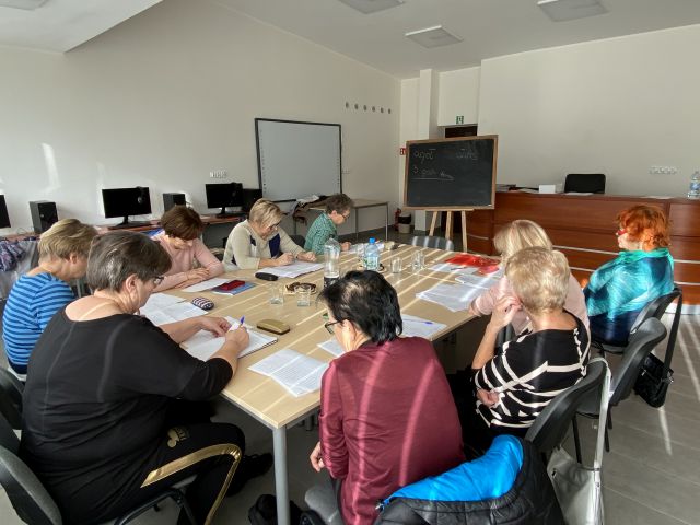 9 seniorek siedzi przy stołach i uczy się języka angielkskiego